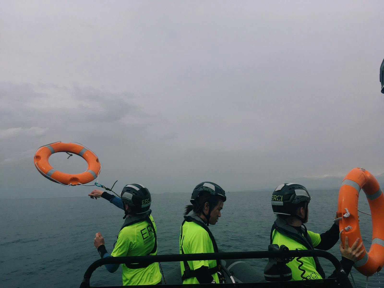 Three search and rescue workers in ERCI t-shirts stand on a boat looking out to sea. One throws a red lifebuoy into the water.