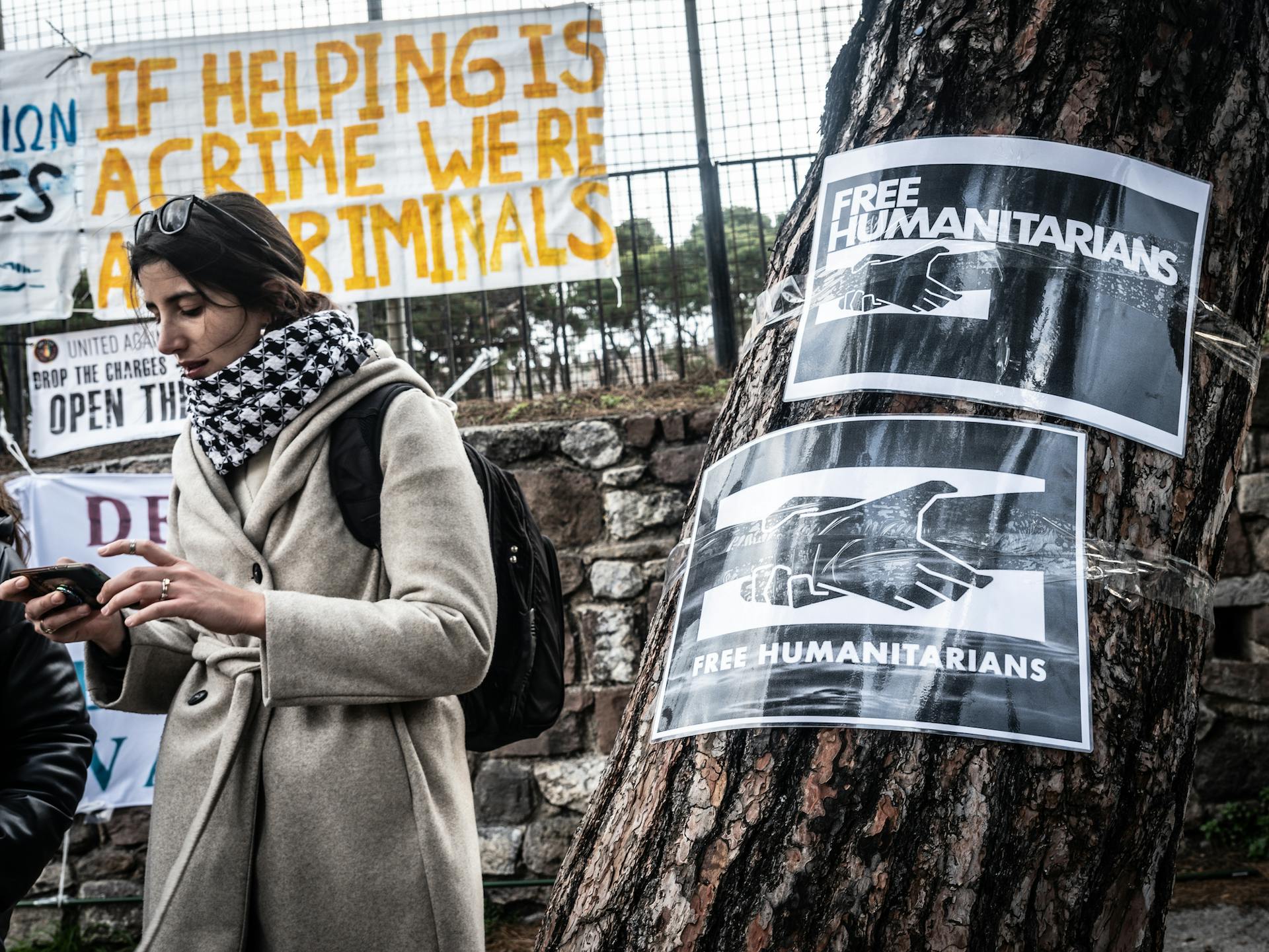 A tree with two 'Free Humanitarians' signs taped to it. Next to it, a person stands in front of a sign that reads 'if helping is a crime we're all criminals.'